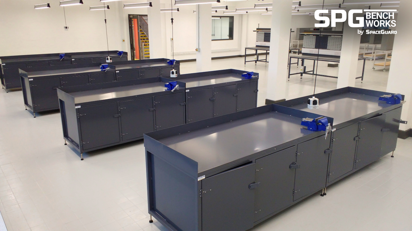 Rows of dark grey heavy duty steel workbenches with cupboards and blue vices in a bright industrial workshop